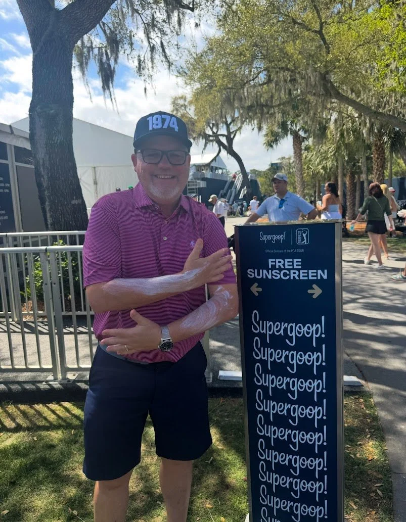 A man standing by a Supergoop branded sunscreen dispenser at THE PLAYERS Championship providing free sunscreen for fans at major outdoor sporting events.