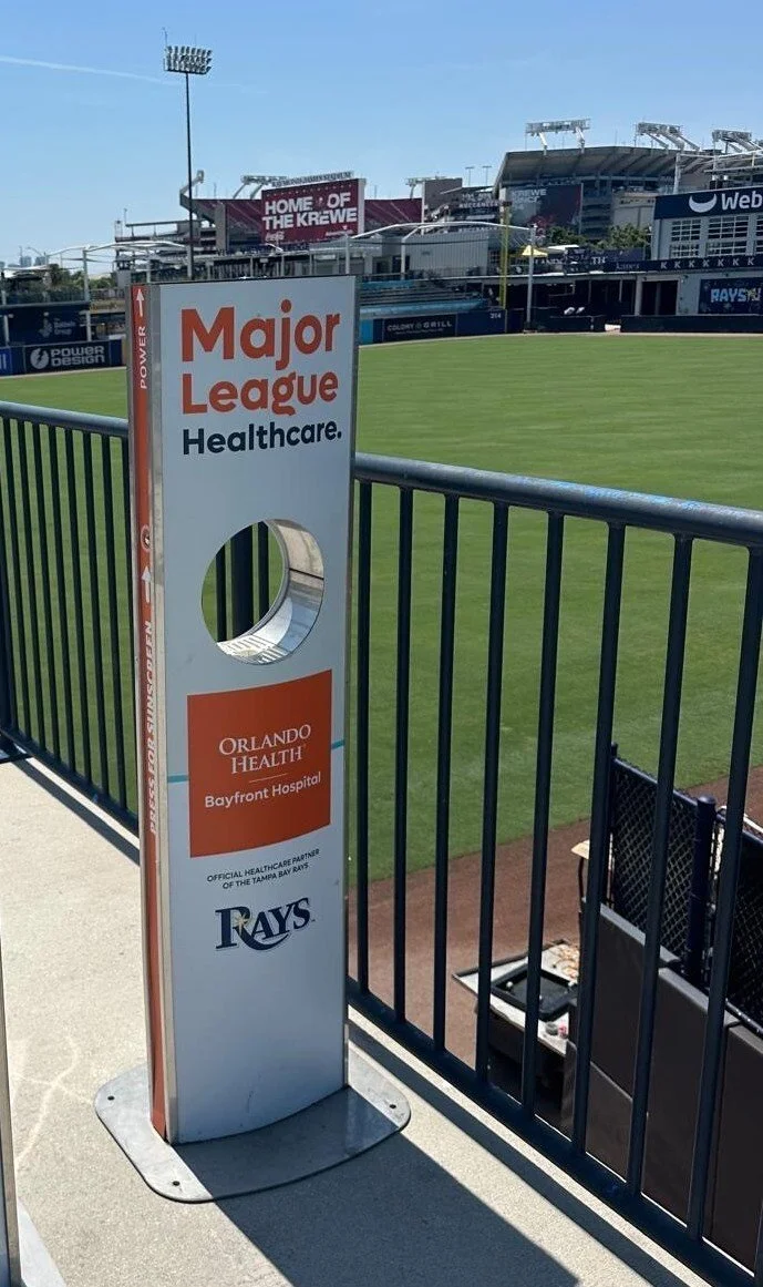Vital Signs Media sunscreen kiosk at Tamps Bay Ray's stadium with empty seats and a field in the background.