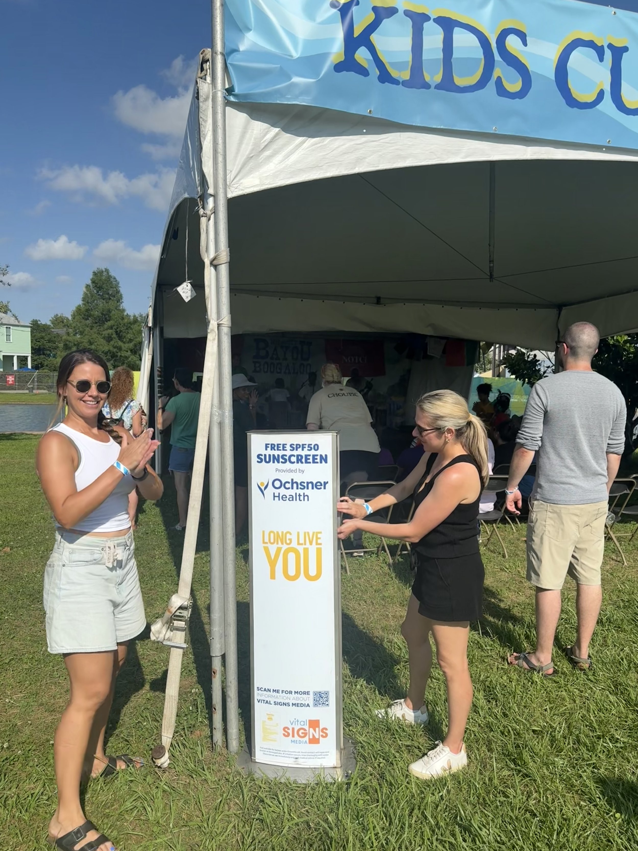People at an outdoor event near a tent with a kiosk offering free SPF50 sunscreen from Ochsner Health.