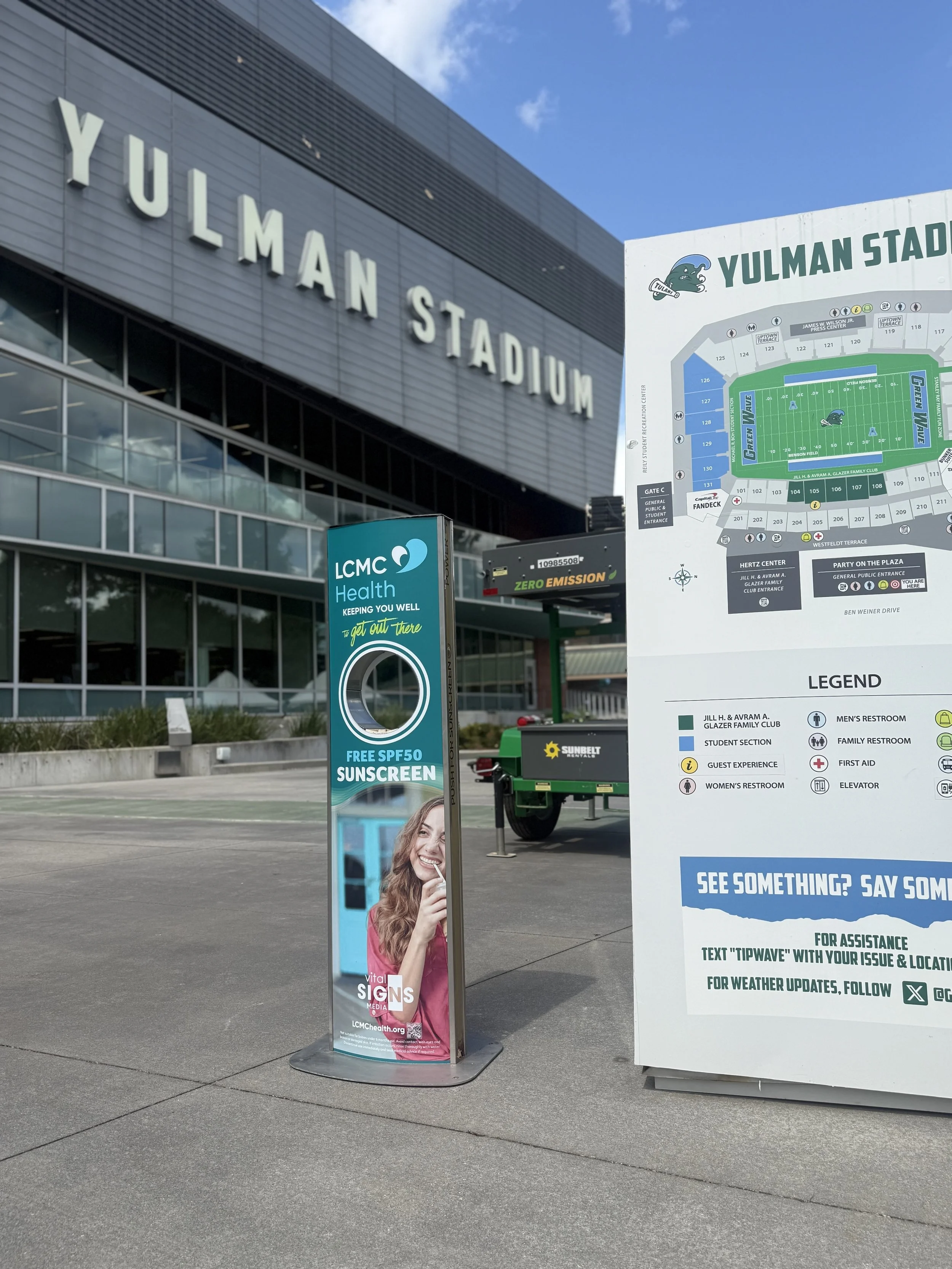 Yulman Stadium exterior with a large sign displaying seating chart and a Nexxus Sunscreen dispenser with a woman smiling on it next to a sunscreen dispenser. 