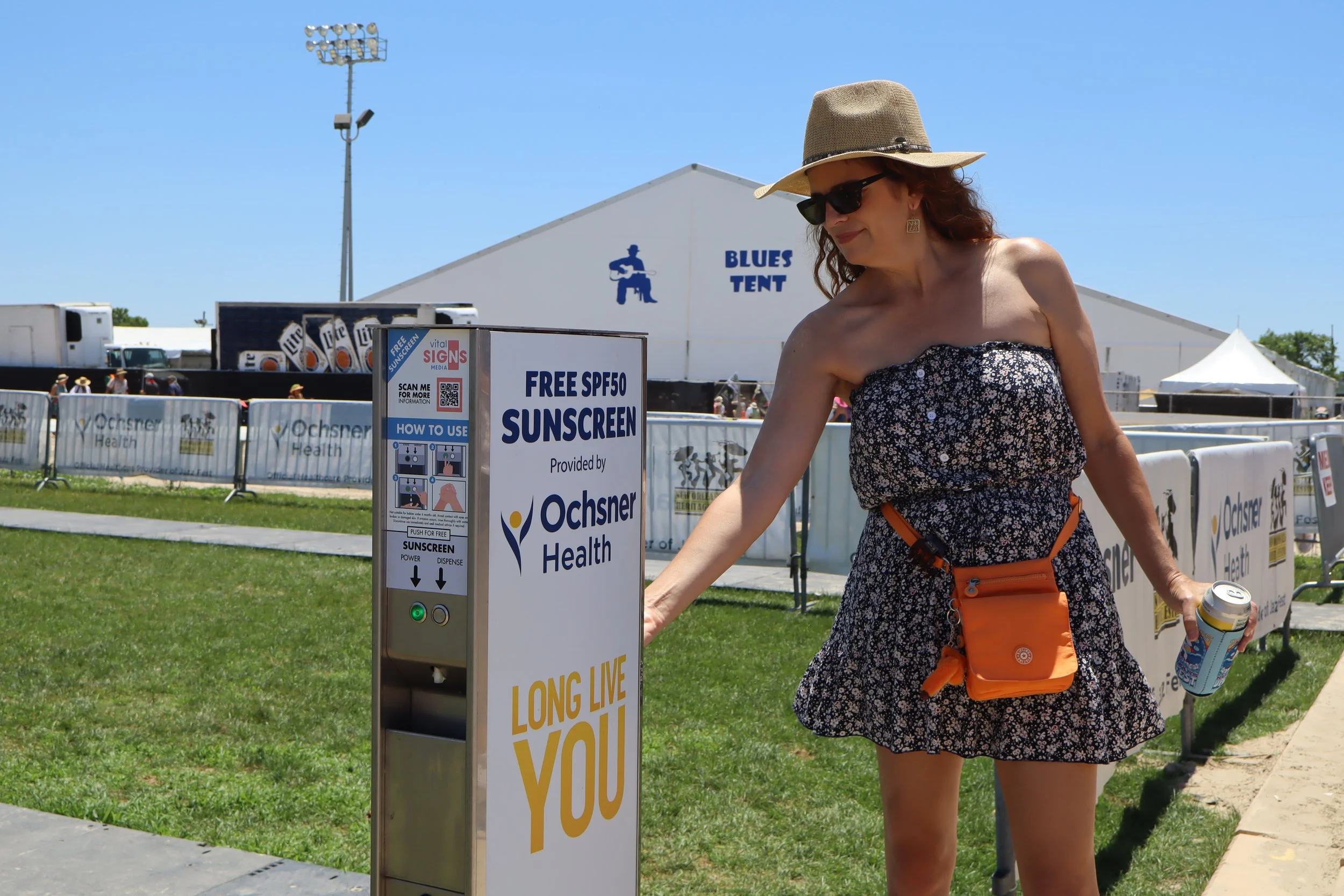 Woman in a floral strapless dress with a tan hat, sunglasses, and an orange crossbody bag, using a free SPF50 sunscreen dispenser at an outdoor event, with tents and a sports field in the background.