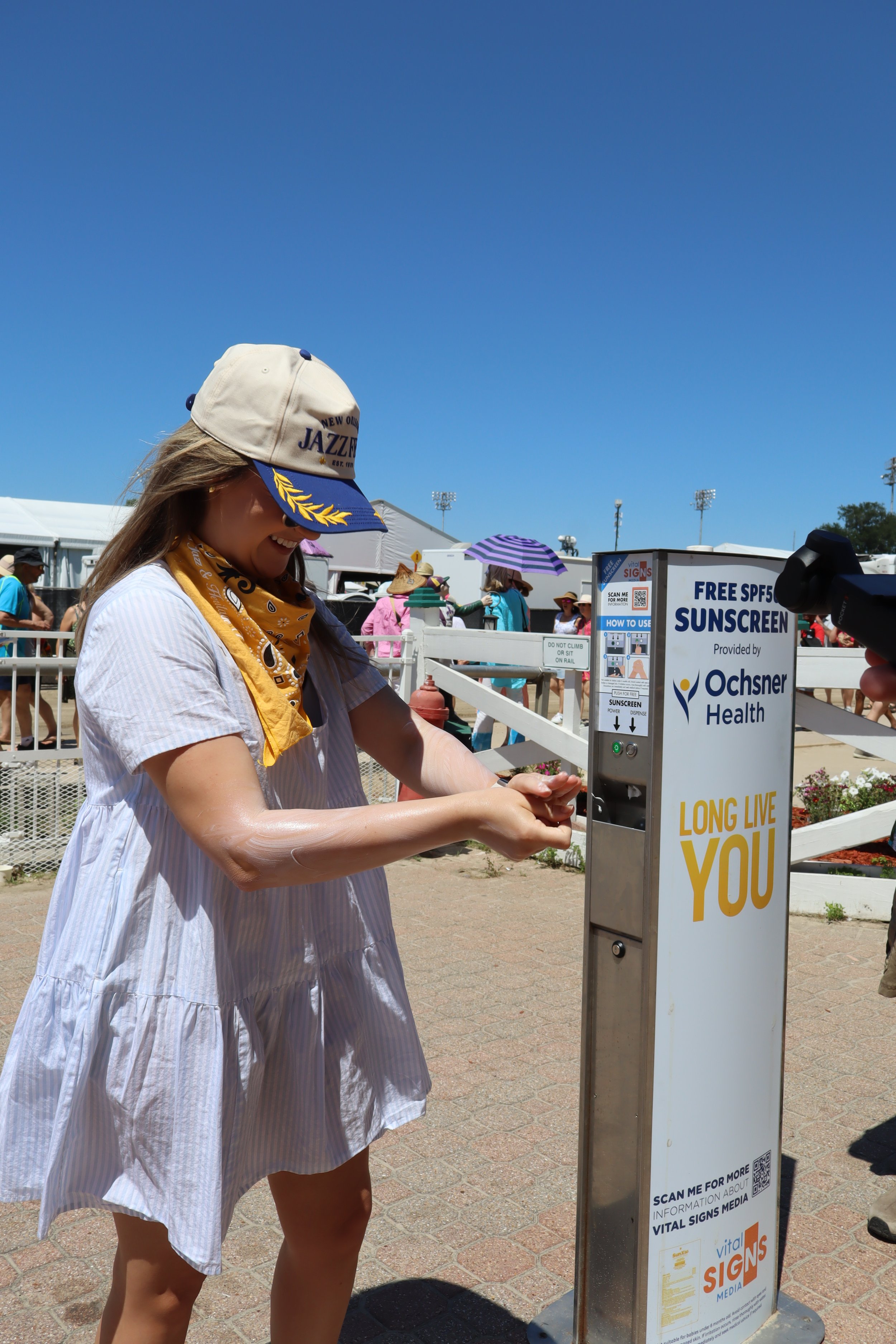 A woman is using a hand sanitizer station at an outdoor event under a bright blue sky, with several people and tents in the background.