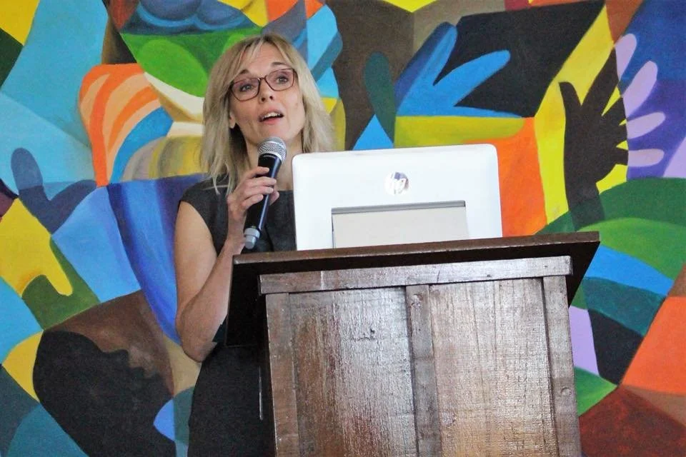Susan Wright speaks confidently into a microphone behind a wooden podium with a computer monitor, in front of a colorful abstract mural.