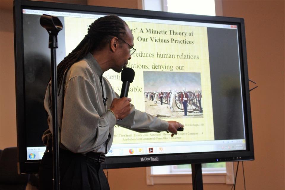 Man with glasses and dreadlocks giving a presentation using a microphone and a touchscreen display, with a slide projected on the screen.