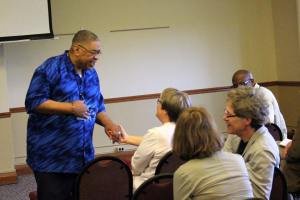 A woman in a blue shirt is speaking to a group of four seated people in a room with beige walls.