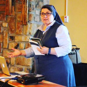 A nun wearing glasses and a traditional habit, holding a notebook, standing in front of a brick wall and a yellow wall. She appears to be giving a presentation or lecture.