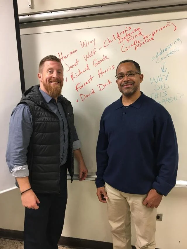 Two men standing in front of a whiteboard with handwritten notes about Harman Wray, Janet Wolf, Richard Goode, Forrest Harris, and David Dark. The board also has notes questioning why and addressing content.