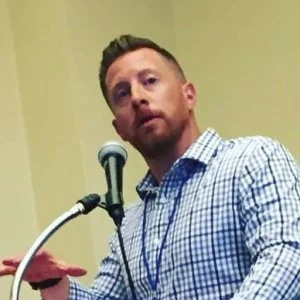 A man with short hair and a beard wearing a checkered shirt speaking into a microphone at a podium.