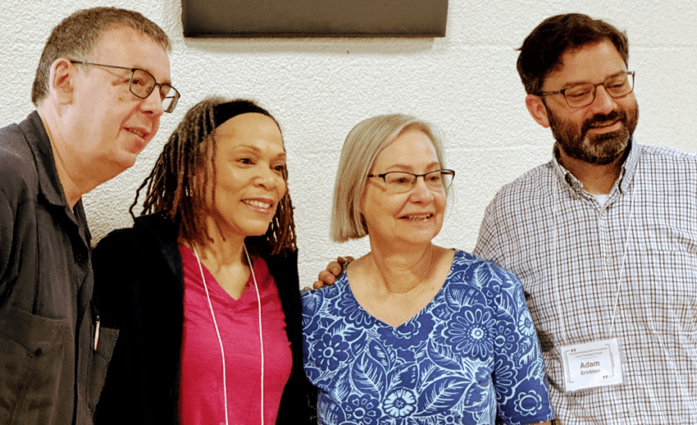 Four people standing together at an indoor event, smiling for a photo. The group includes two men and two women, all wearing glasses.