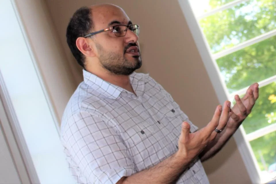 A man with glasses and a beard, wearing a checkered shirt, standing indoors with a window behind him, gesturing with both hands as if explaining something.