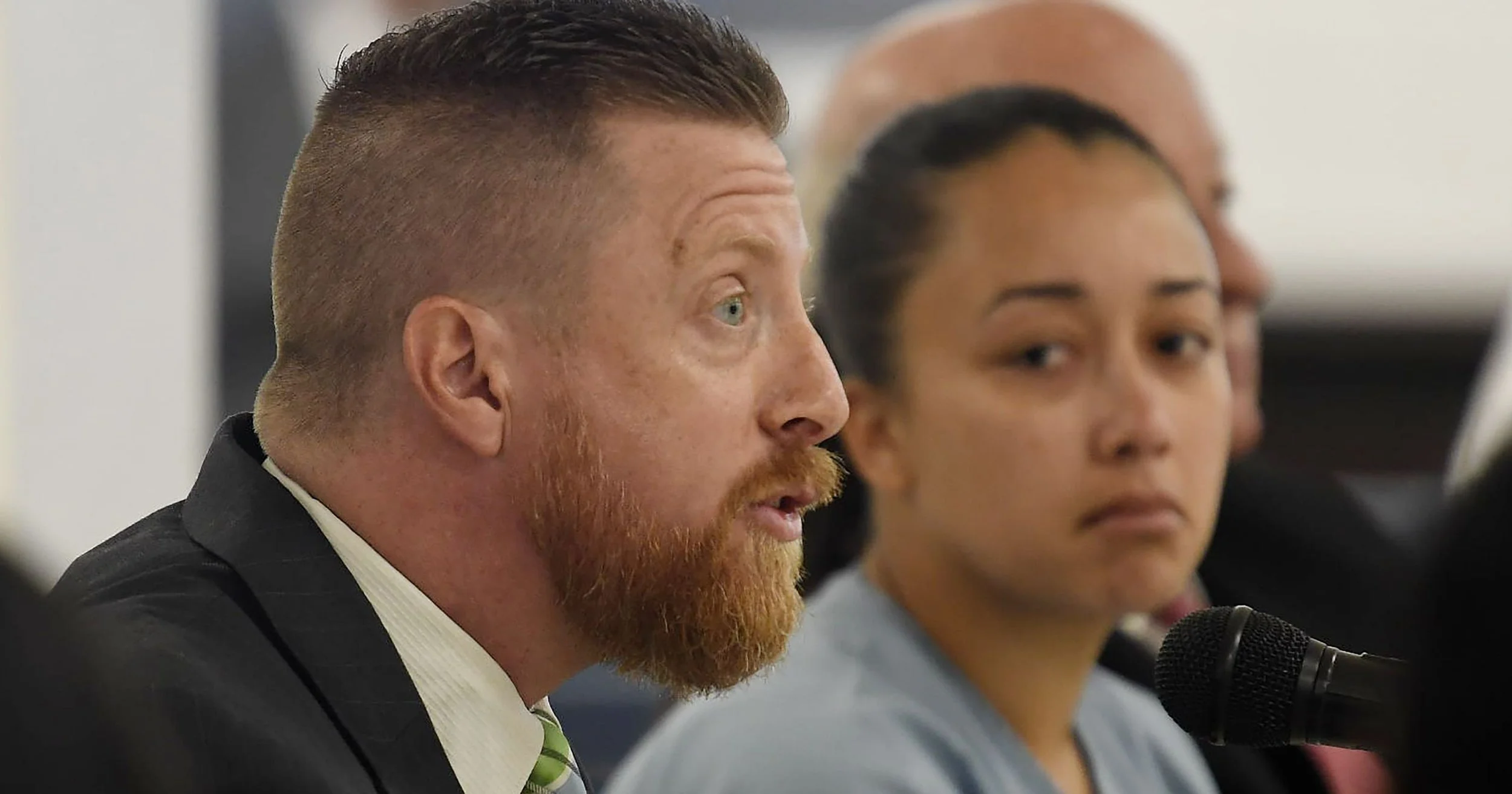 A man with a red beard and short brown hair speaks into a microphone during a meeting, seated next to a woman with dark hair tied back, while other people in the background listen attentively.