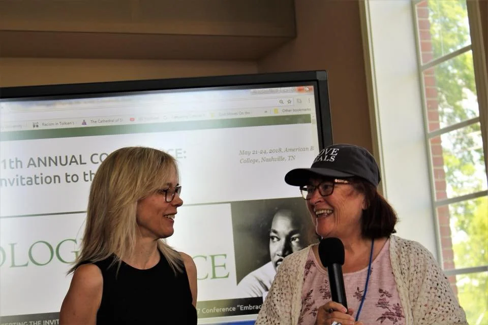Two women smiling and talking, one holding a microphone, in front of a large screen with event information, near a window with greenery outside.