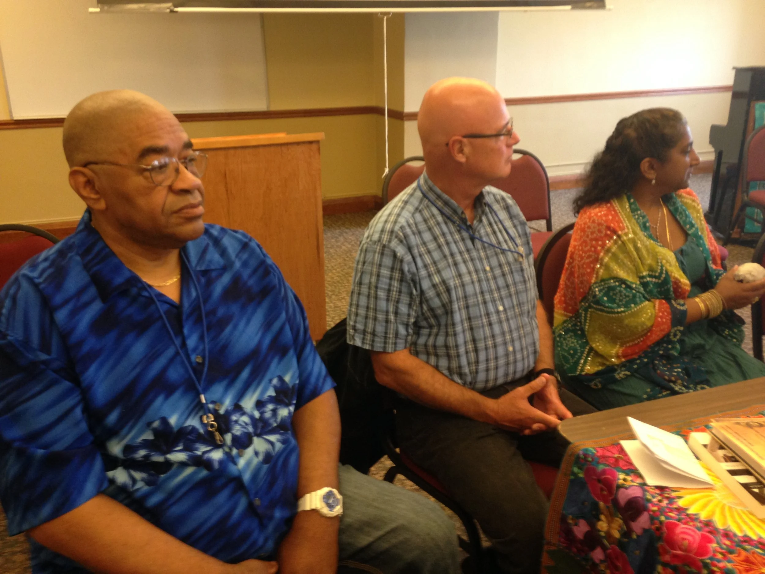 Three people sitting side by side in a meeting or conference room. The first person, on the left, is an older man with glasses and a blue patterned shirt. The middle person is a bald man wearing glasses and a plaid shirt. The third person, on the rig