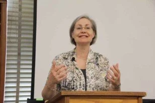 Older woman with short gray hair giving a speech at a podium, smiling, wearing a patterned blouse in a classroom or conference room.