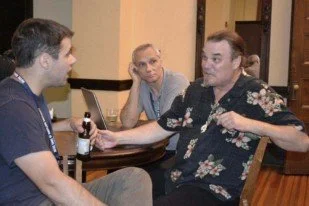 Three men sitting at a table, engaged in conversation. One man is wearing a black floral shirt and gesturing with his hands. The other two men are listening; one has short dark hair, and the other has lighter hair and is resting his face on his hand.