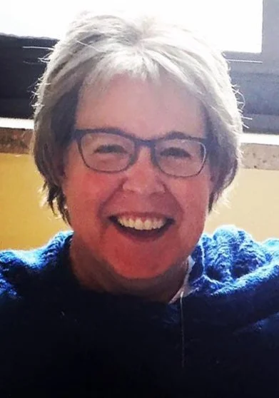 A smiling woman with short gray hair, glasses, and a blue sweater.
