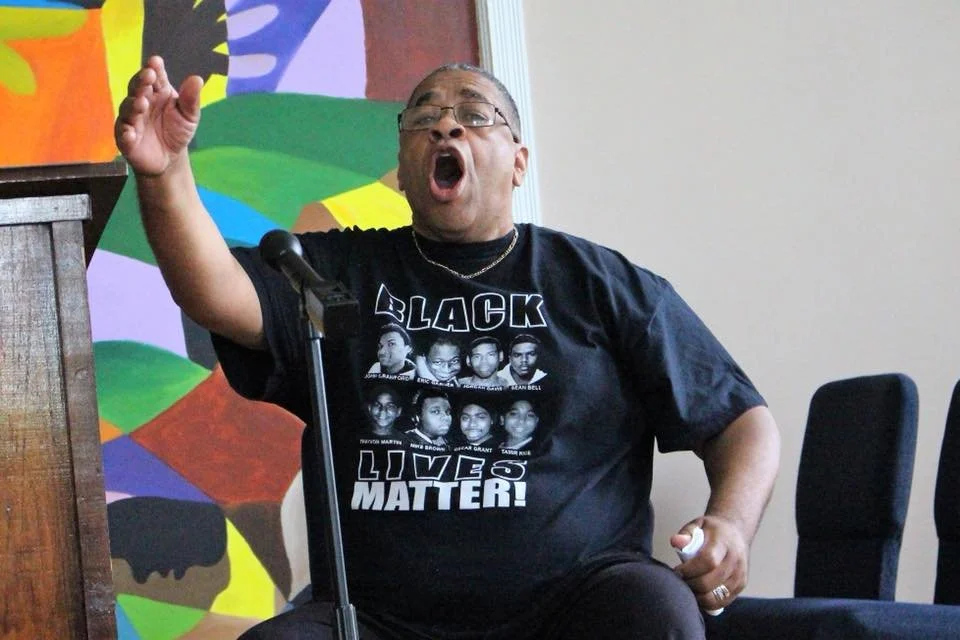 A middle-aged woman wearing glasses and a black T-shirt with photos of Black individuals and the words 'BLACK LIVES MATTER' is speaking passionately at a microphone, with her mouth open and one arm raised.