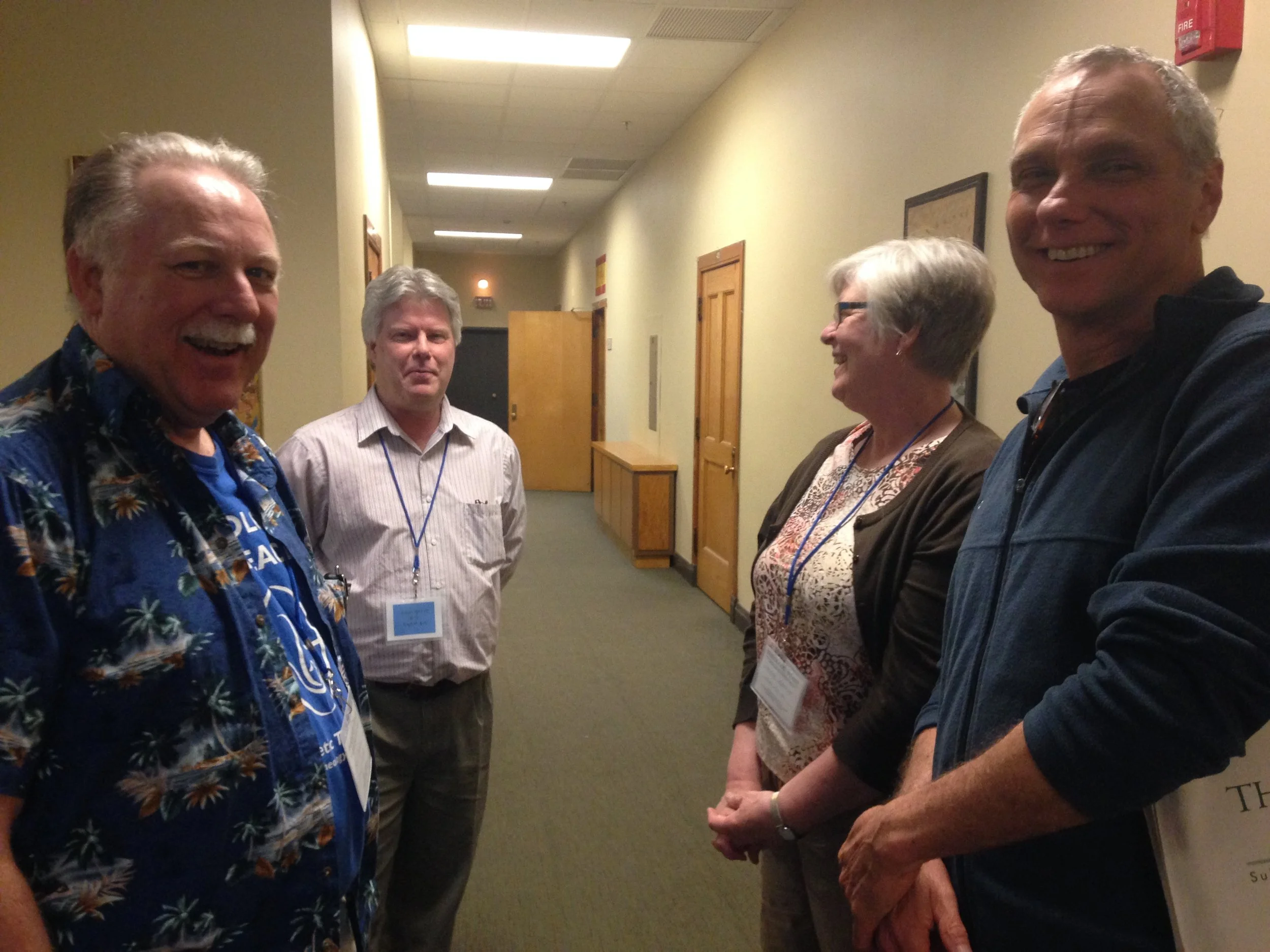 A group of five people in conversation in an indoor hallway with wooden doors and a fire alarm on the wall.