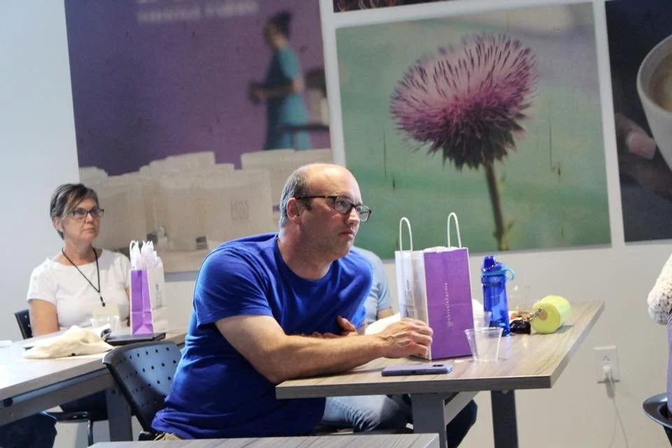 Shannon Mullen and other conference participants listen attentively to a presentation at Thistle Farms, with purple gift bags beside them and large posters of flowers on the wall behind them.
