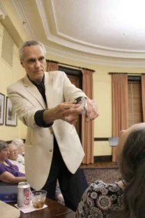 A man in a white jacket performing a card trick for a seated audience in a decorated room.