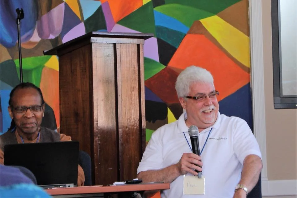 Two men, one with gray hair and glasses holding a microphone and smiling, and another with glasses and dreadlocks sitting behind a laptop, in a colorful room with a geometric mural.