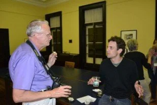 Two men having a conversation at a table, one holding a drink and a camera around his neck, the other gesturing with his hands.