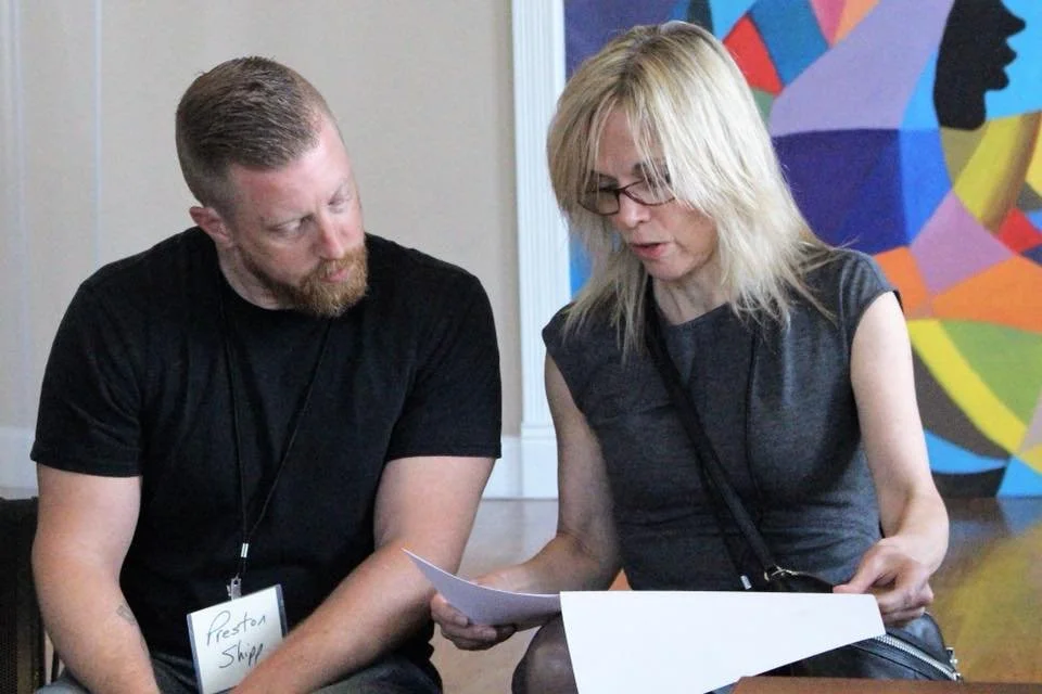 Aman and woman sitting closely together looking at a piece of paper. The man has a beard and short hair, wearing a black shirt. The woman has blond hair, glasses, and is wearing a dark gray top, also with a badge around her neck. They are in an indoo