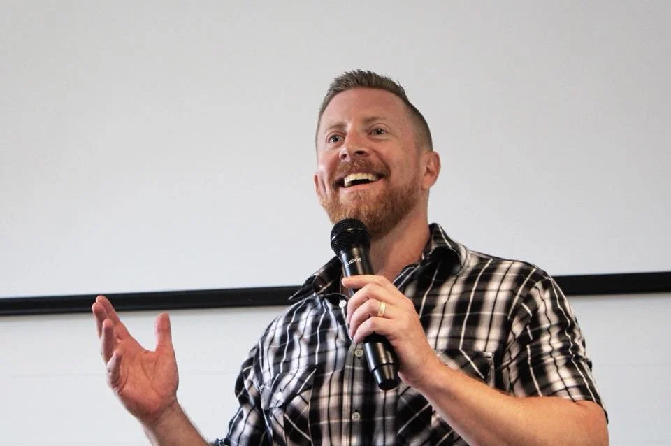 A smiling Preston Shipp holds a microphone in his left hand and gestures with his right hand during a presentation.