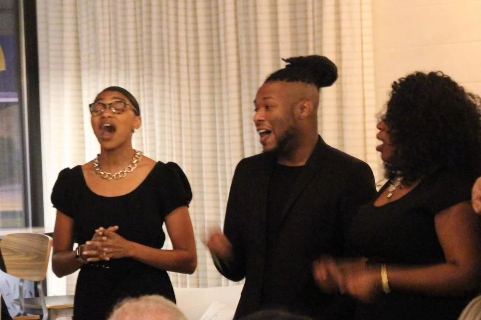 Three people at a social gathering, two women and a man, appear to be singing or talking happily, standing indoors with beige curtains in the background.