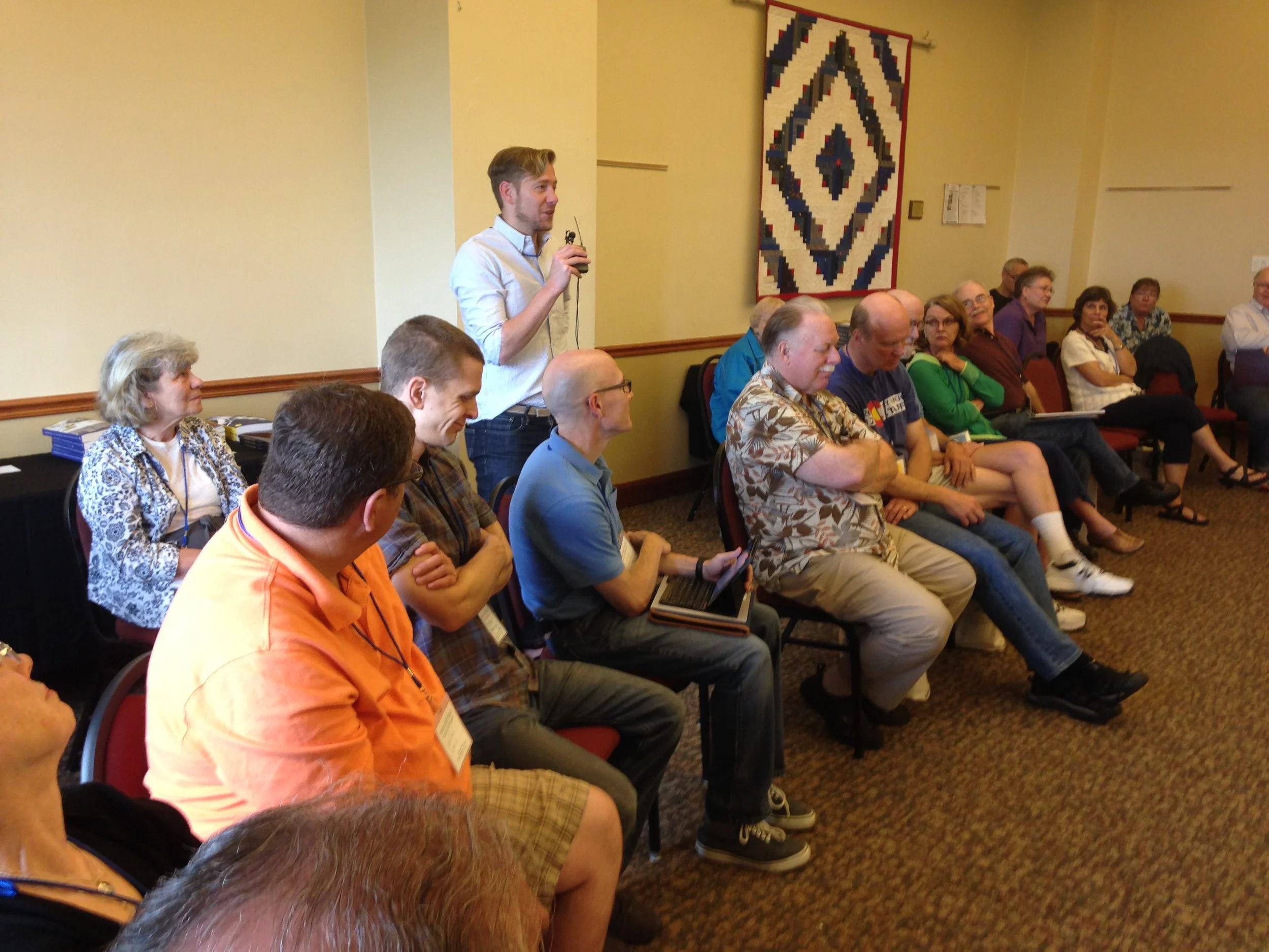 A man holding a microphone appears to be speaking or giving a presentation in a room with several seated audience members. The room features a decorative textile hanging on the wall and a table with books and papers.