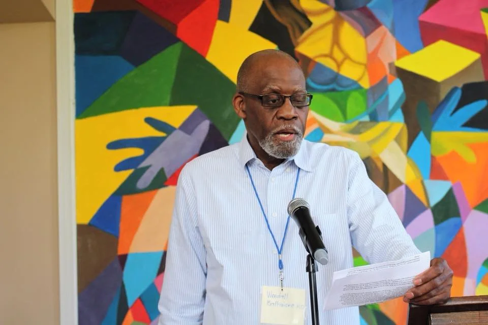 An elderly man with glasses and a beard is speaking into a microphone while holding papers, standing in front of a colorful abstract mural.