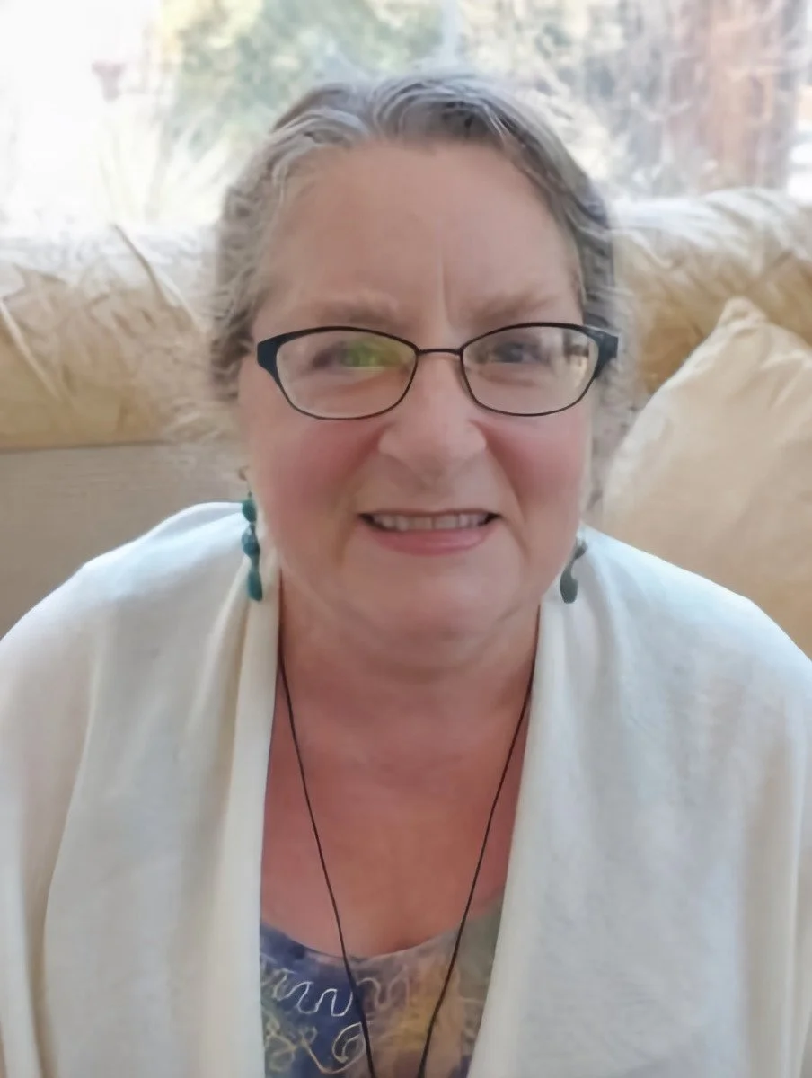 A senior woman wearing glasses, earrings, and a necklace, sitting on a beige couch on a sunny day with a window behind her.