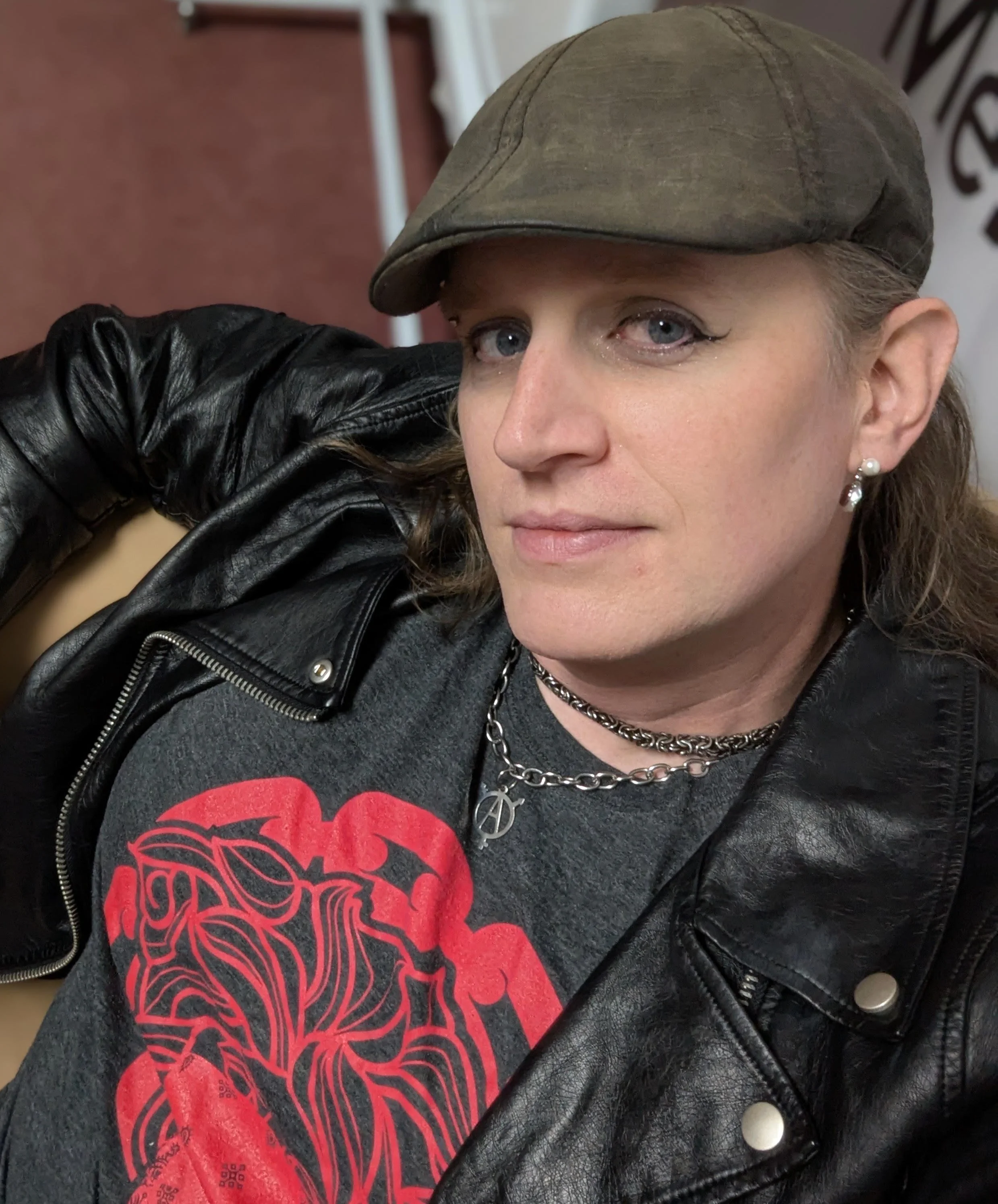 Billie Hoard, co-author of Eucontamination. A white woman wearing eyeliner, a newsboy cap, a grey T-shirt with a vivid red screen print, and a black leather bomber jacket.
