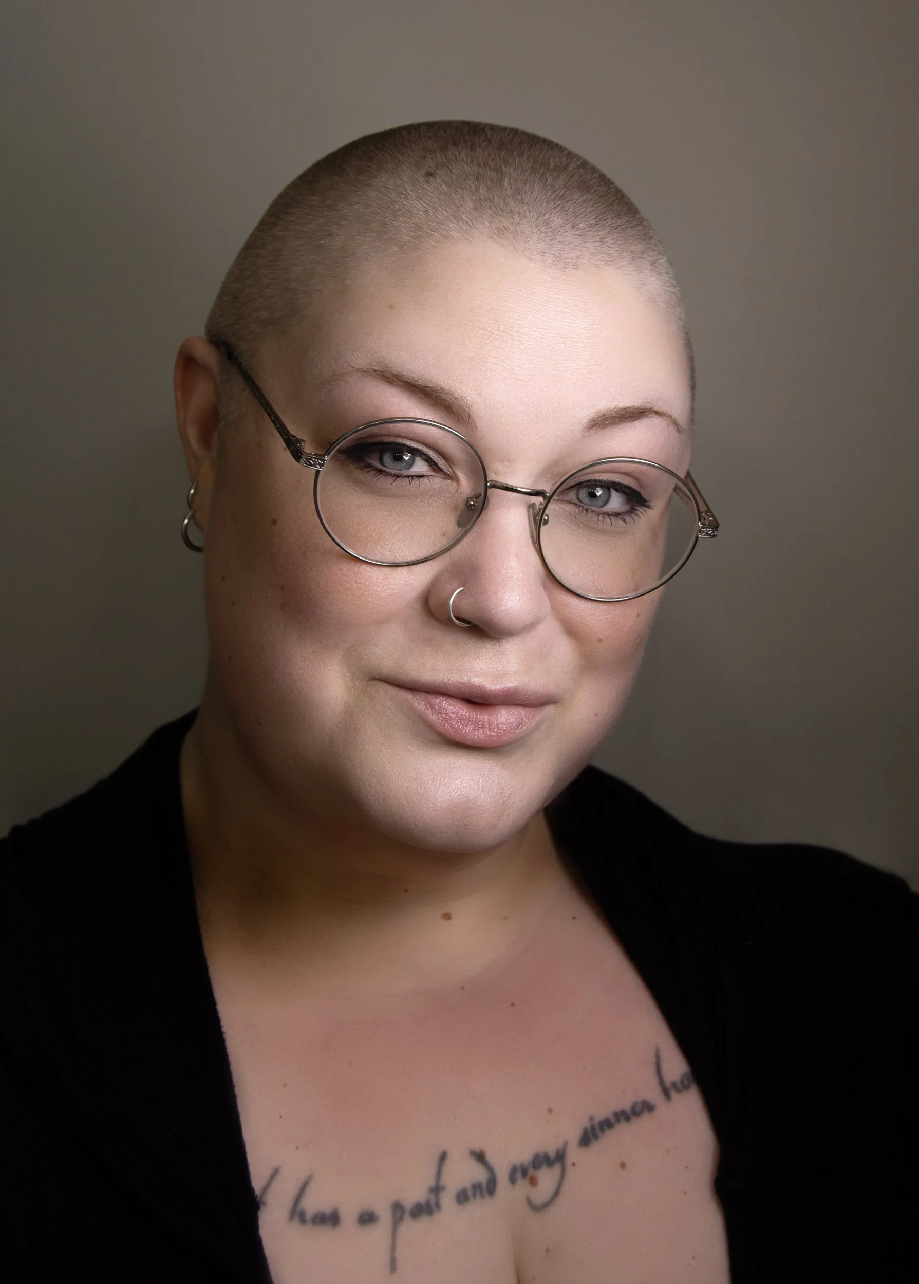 Close-up portrait of a woman with a shaved head, wearing round glasses, earrings, and a nose ring. She has light skin, blue eyes, and a tattoo on her chest with cursive writing.