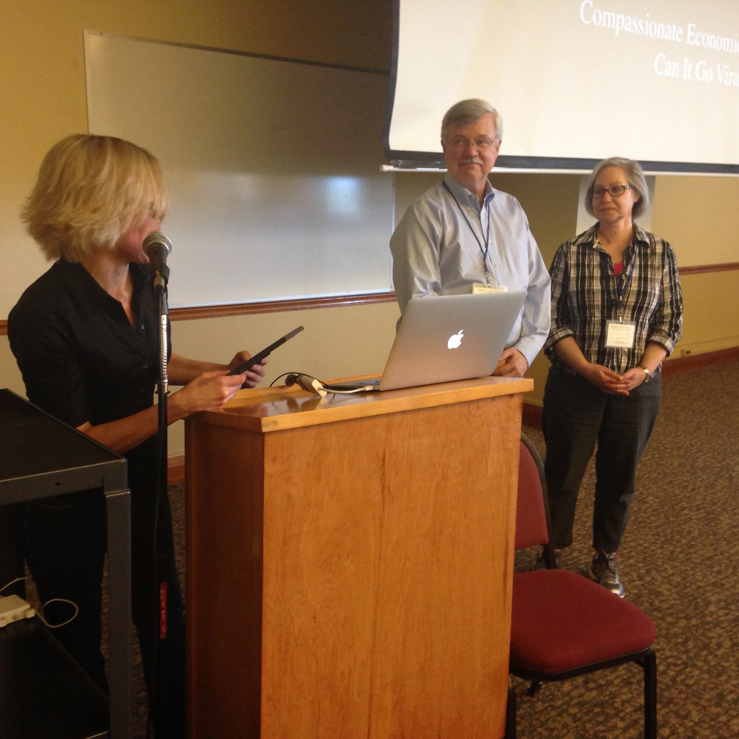 A woman with blonde hair speaking into a microphone with a laptop, and two people standing in front of a large screen at a conference or presentation. One man and one woman, both wearing conference badges, are looking toward the woman at the podium.