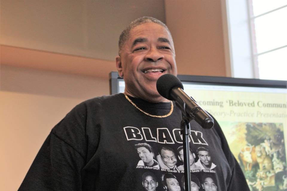 A man speaking into a microphone, wearing a black T-shirt with images of historical figures on it, in an indoor setting with a presentation screen in the background.
