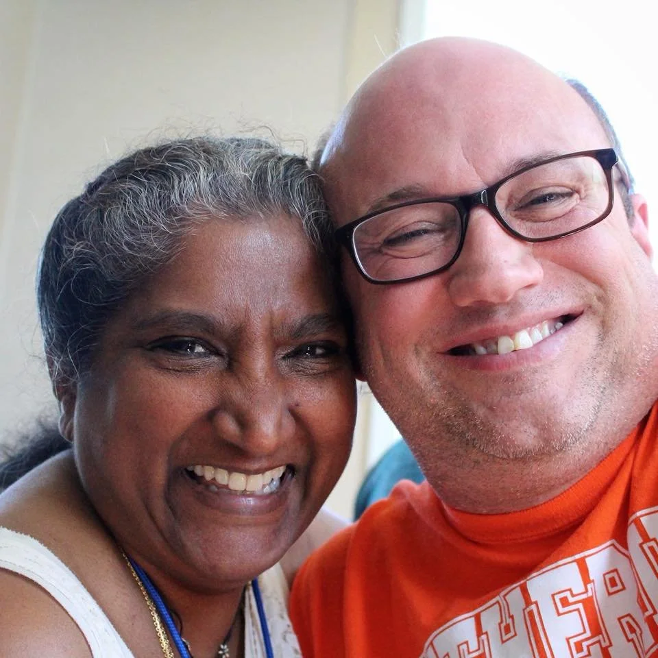 A smiling woman with gray hair and a man with glasses wearing an orange shirt, both smiling and close together for the photo.