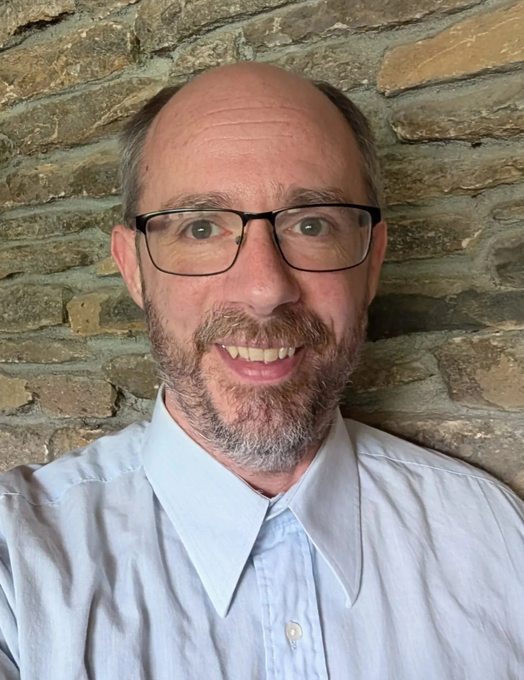 A smiling man with glasses, wearing a light-colored collared shirt, against a stone wall background.