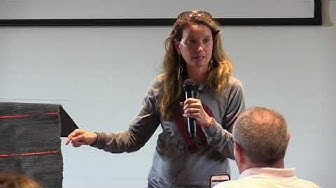 A woman speaking into a microphone at an indoor event, facing an audience member with a bald head.