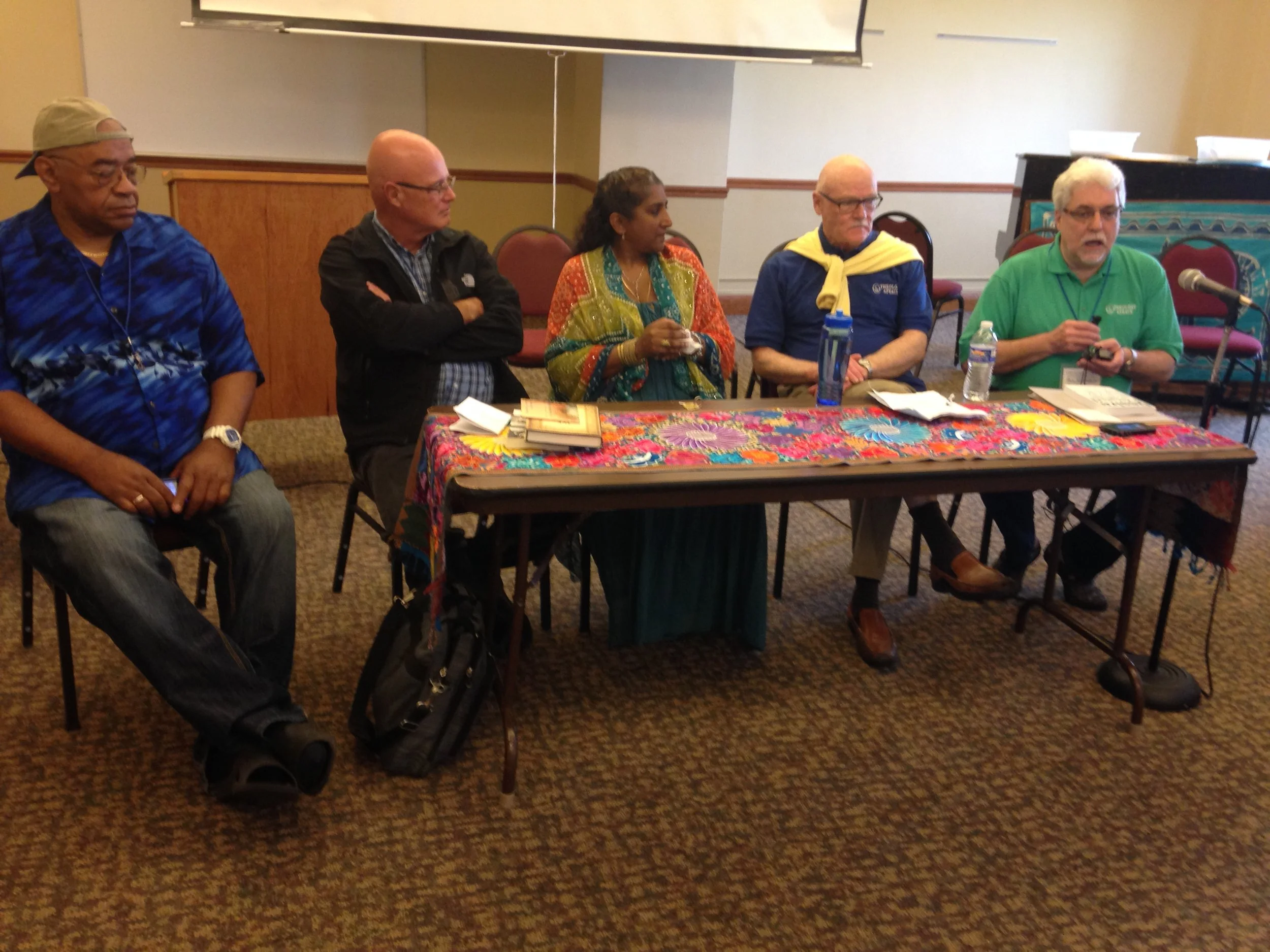 Five people sitting at a table with colorful tablecloth, listening to a man in a green shirt who has a microphone in front of him. The table has books, a water bottle, papers, and a microphone. The background shows a room with chairs and a projection