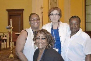 Group of four women smiling together indoors, with a candle holder and wooden furniture in the background.