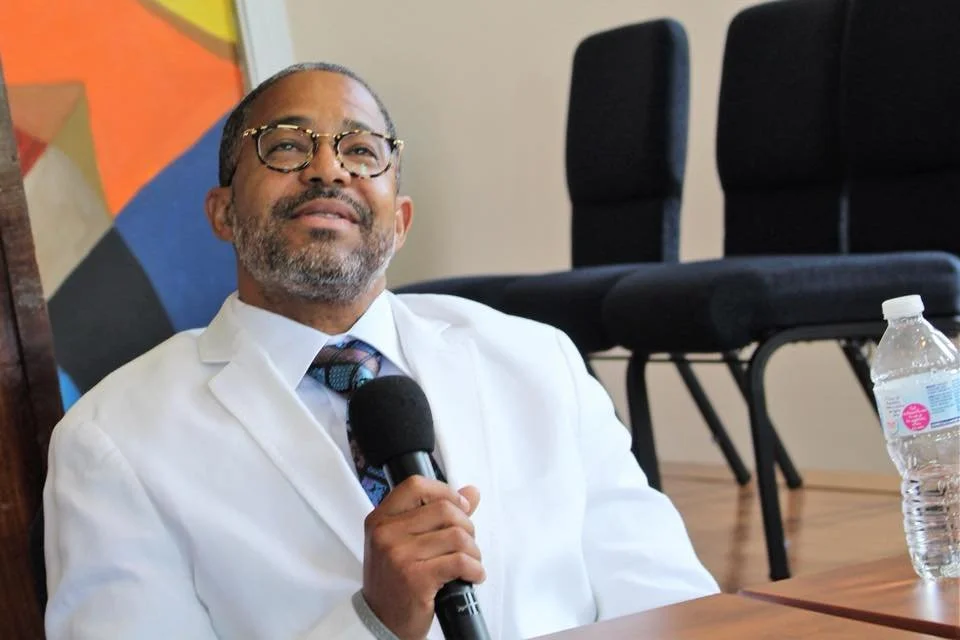A smiling man in a white shirt and glasses holding a microphone, sitting in a conference room with chairs and a water bottle on the table.