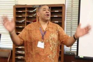 Man speaking or giving a presentation in a classroom or office, gesturing with hands raised, wearing an orange shirt.