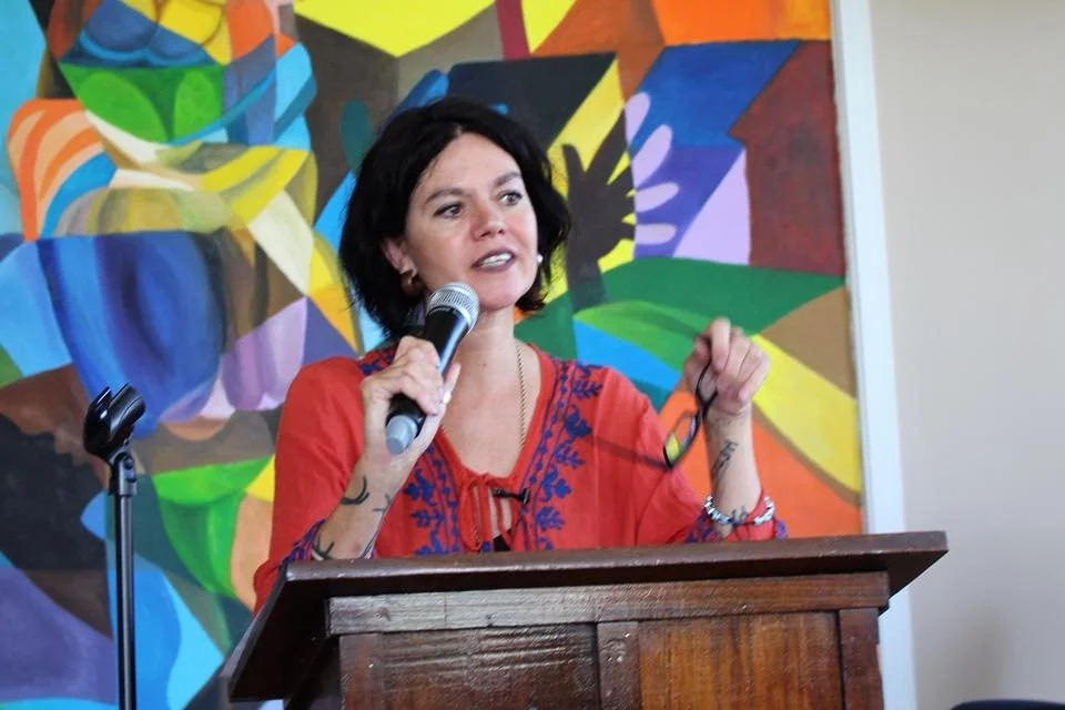 A woman with black hair, wearing a red embroidered top, speaking into a microphone behind a wooden podium, with a colorful abstract mural in the background.