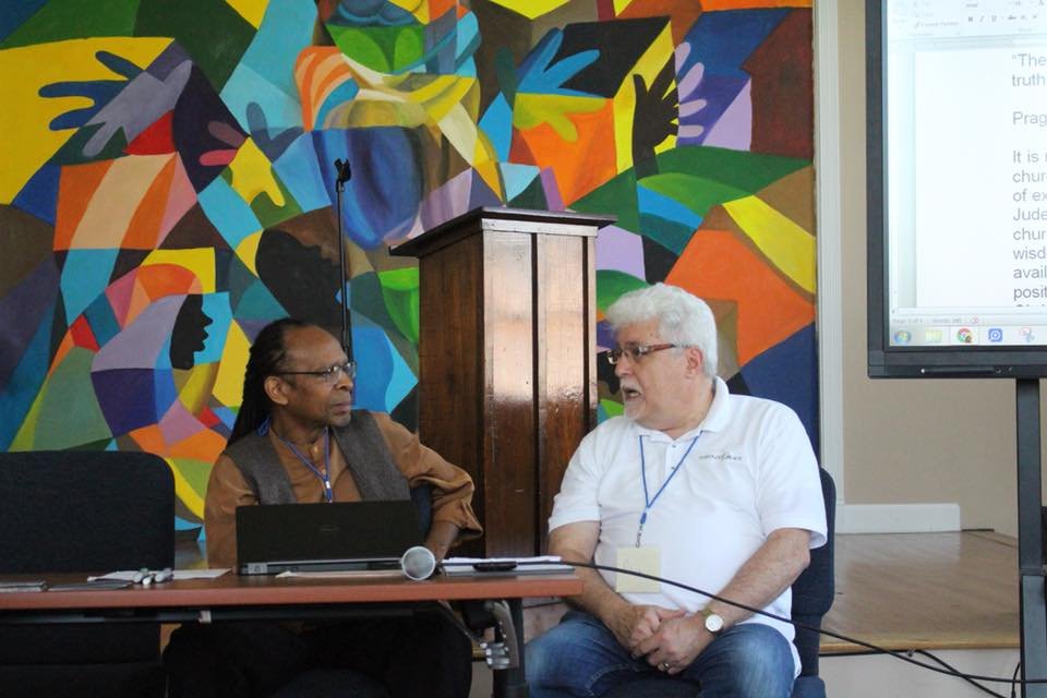 Two men seated at a table having a conversation in a room with a colorful abstract mural on the wall and a large screen displaying text.