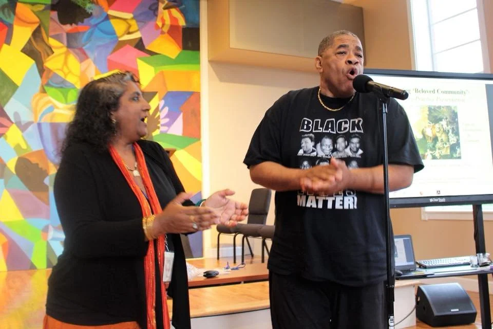 Patty Prasada-Rao and Elder C.W. Harris lead the conference participants in song, with a colorful mural in the background and a presentation slide visible on a screen.