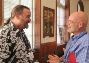 Two men smiling and shaking hands inside a room with wooden paneling and framed art on the walls.