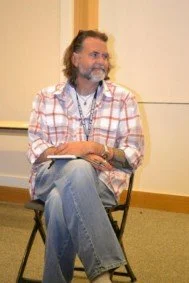 A mature man with glasses, a beard, and long hair, sitting on a chair, smiling, wearing a plaid shirt and jeans, holding a book. He is Richard Beck, author of Unclean: Meditations on Purity, Hospitality, and Mortality.