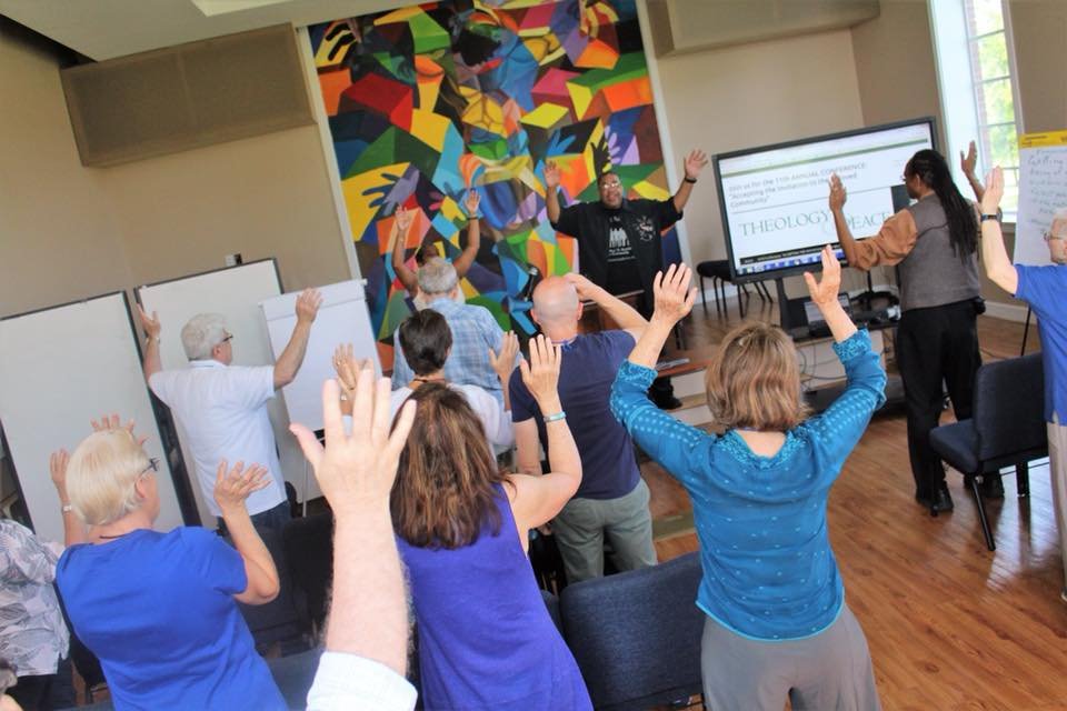Patty Prasada-Rao and Elder C.W. Harris lead conference participants in song, with a colorful mural on the wall and a large screen displaying presentation slides.
