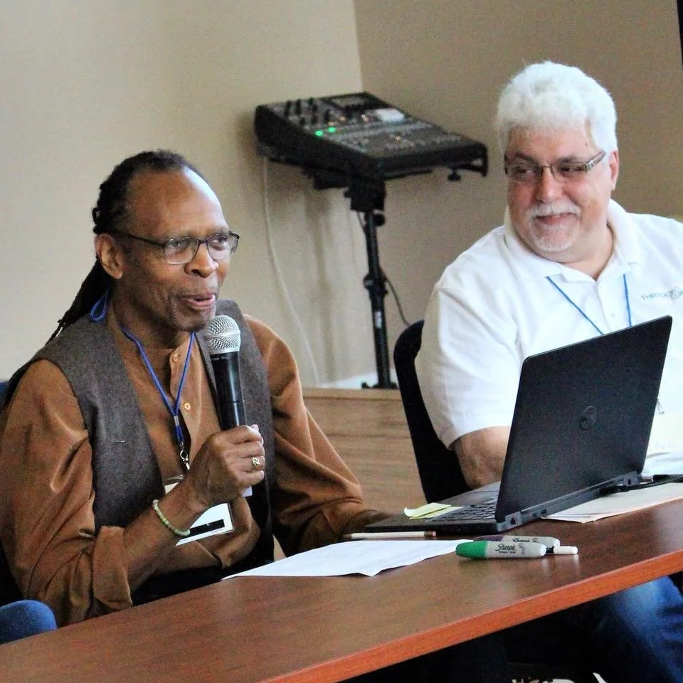 Rev. Thee Smith speaks into a microphone while Paul Nuechterlein looks on. Both men are sitting behind a table and facing an audience. There is a laptop on the table in front of them.